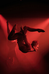 Female aerialist performs a dramatic backbend pose in mid air using a harness, arching her body with bent legs under red spotlight for an intense stage effect. © Dima