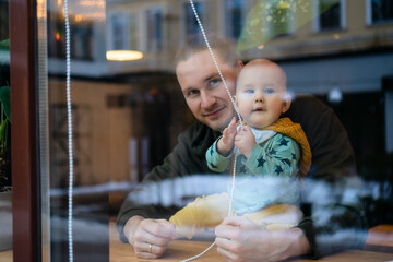 Young father with toddler in cafe holding child, visible through window. Family values, parenting love, bonding and care in public place with warm atmosphere and happiness.