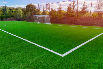 A football field on a beautiful summer evening, with the sun setting and a blue sky overhead. The green grass is dotted with white lines marking the penalty area and the goal posts. In the distance, a