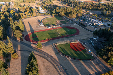 Aerial drone picture of college sports fields in Oregon City with soccer, baseball and track areas, students practicing on green lawns during fall sunset, surrounded by trees and campus buildings