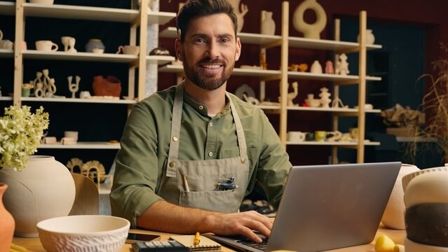 Happy smiling Caucasian man guy small business owner businessman successful entrepreneur crock potter artist male working computer typing laptop internet website looking camera inside pottery workshop - Powered by Adobe