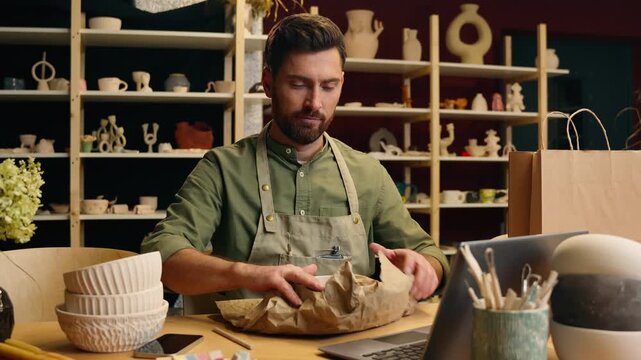 Careful Caucasian man guy businessman male small business owner potter salesman crock packing fragile clay bowl artwork pottery ceramic in shopping bag package typing laptop delivering indoor workshop