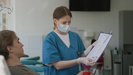 Masked nurse in blue scrubs asking young man to sign documents before donating his blood in clinic