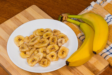 Dried banana chips on plate and two fresh unpeeled bananas