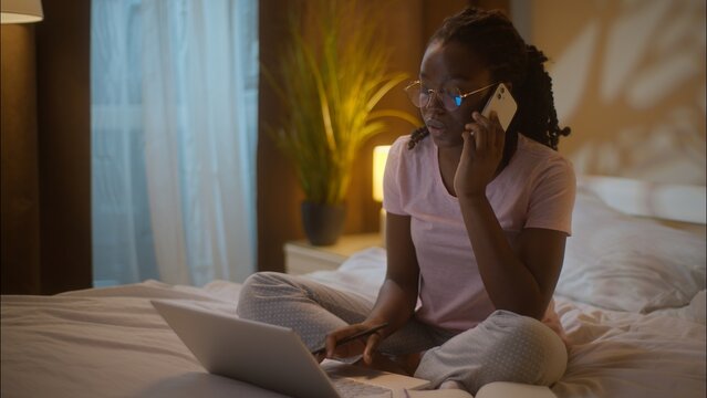 Multitasking African American business woman student female girl businesswoman in glasses talk mobile phone call working laptop freelance typing computer writing notes sitting bed at home late evening