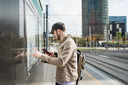 Young male student buying transport pass at urban tram train station using mobile phone app and ticket machine