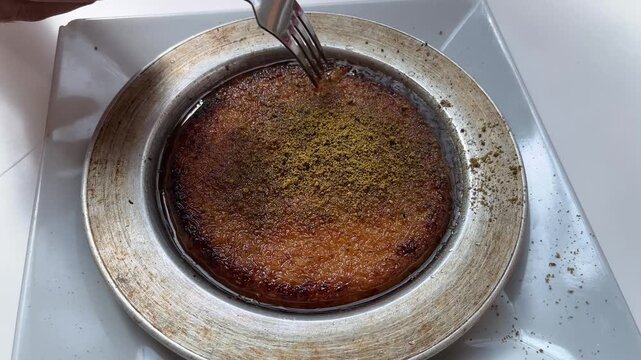 Person taking a slice of traditional Turkish kunefe dessert with fork from metal plate topped with ground pistachio