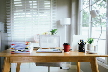 A bright modern office with a wooden desk, laptop, stacked documents, and natural light from large windows.