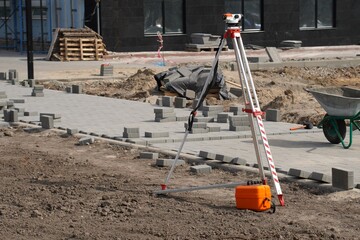 Surveying equipment positioned on an active construction site with paving in progress. Bricks are laid out on prepared ground, indicating organized planning and development.