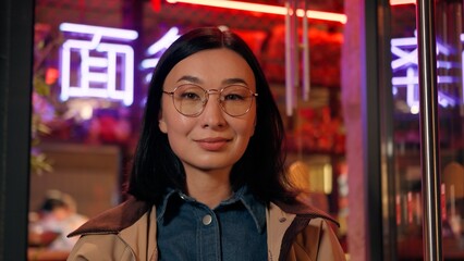 Obraz premium Smiling happy Japanese Korean glad business woman female client businesswoman in glasses looking camera smile outdoors night city neon lights. The inscription noodle in Chinese. Translation: 'noodle'.