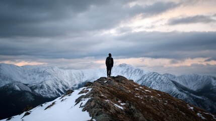 Lone backpacker stands on hilltop, overlooking vast alpine landscape with snow capped mountains under dramatic, cloudy sky. scene evokes sense of adventure and solitude