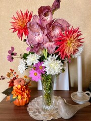 A colorful bouquet featuring dahlias, gladiolus, and chrysanthemums in a glass vase. Autumn decorations include a small pumpkin and textured bird figurine on a wooden surface.