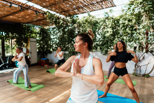 Group of women actively participating in a yoga class. Performing a gentle stretching pose in a bright studio surrounded by lush green plants. Focusing on wellness. Mindfulness. And healthy lifestyle