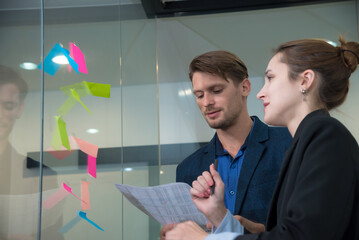 Creative professionals collaborate in modern office setting, discussing ideas while reviewing documents. Colorful sticky notes on glass wall enhance innovative atmosphere