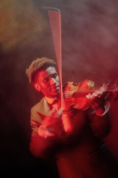 Black young adult man playing violin on stage, wearing suit, captured in motion with blurred effect, focused expression, dramatic lighting, performing classical music