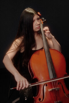 Hispanic girl playing cello with focused expression, long dark hair framing face, holding bow in right hand and pressing strings with left hand on stage under spotlight
