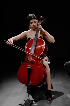 Young adult Hispanic woman playing cello on stage, sitting and focusing on performance, holding bow and instrument with both hands, dark background emphasizing musician and cello