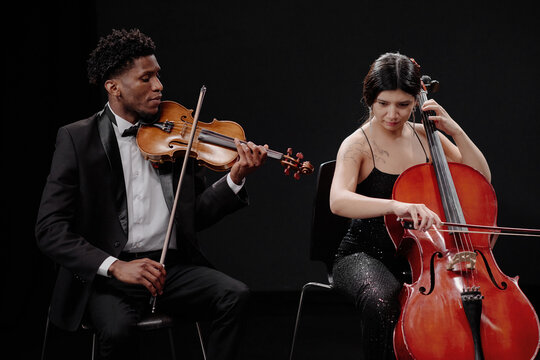 Young adult Black man playing violin and young adult Hispanic woman playing cello sitting side by side on stage performing classical music together against dark background
