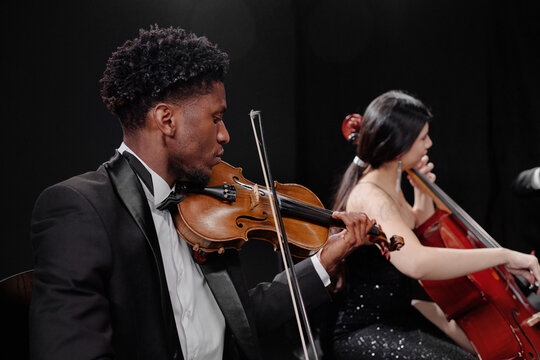 Black young adult man playing violin and Hispanic young adult woman playing cello on stage, both wearing formal attire, focused on performing classical music in concert setting