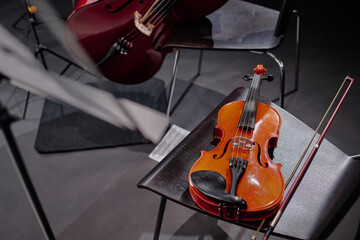 Violin resting on chair with bow placed beside instrument, cello partially visible in background, empty chairs arranged on stage suggesting preparation for classical music performance
