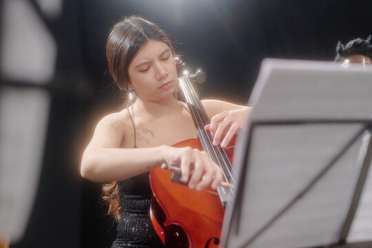 Young adult woman playing cello with focused expression, reading sheet music during performance, long dark hair visible, seated on stage in professional music setting