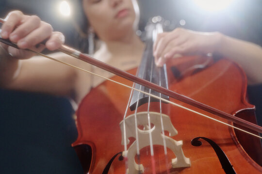 Young adult woman playing cello on stage, focusing on hand positioning and bow technique, close up of instrument and fingers highlighting musical performance and concentration