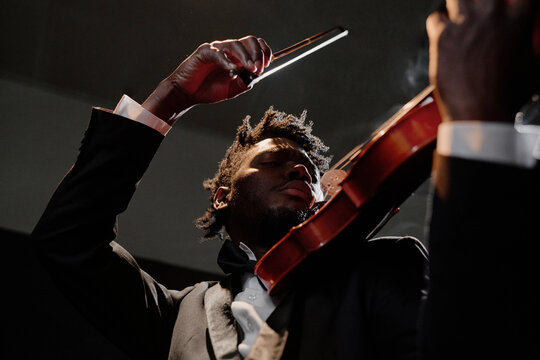 Black young adult man playing violin on stage, wearing formal suit, holding bow above strings, focusing intently on performance, dramatic upward angle emphasizing concentration