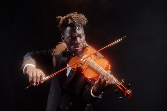 Black young adult man playing violin on stage, wearing formal suit, focusing intently on performance, dramatic lighting highlighting expressive face and hands, dark background
