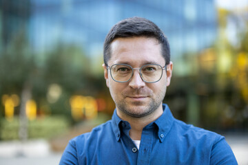 Obraz premium Entrepreneurial man wearing a blue shirt and glasses looking directly at the camera with a confident expression, representing professionalism and success in an urban setting