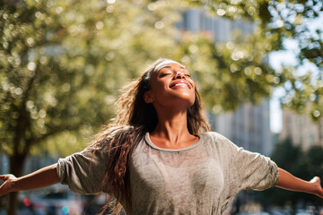 Young woman with arms outstretched wearing gray loose t-shirt and blue jeans in urban park with green trees and buildings in background under golden sunlight