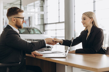 A bearded man in a suit, a manager, a car salesman in a car dealership, who signs a contract at the table with a woman buyer and shakes her hand