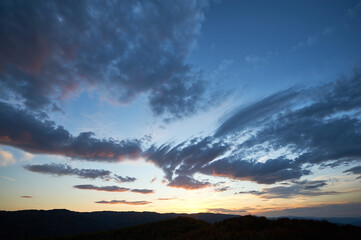 Fototapeta premium Stunning sunset fills sky with rich hues of blue and orange. Dramatic clouds with hints of pink and gold spread across sky. Silhouettes of hills in foreground add depth to breathtaking evening scene.