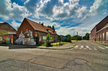 Wide-angle street view of abandoned Belgian village showing boarded houses with colorful graffiti under dramatic cloudy sky. Urban exploration photography capturing rural decay.