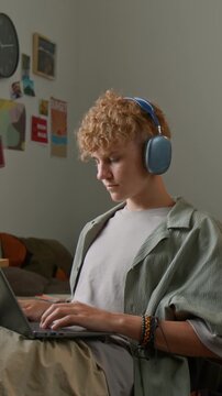 Vertical shot of teenage boy using computer at home and typing on keyboard while sitting at desk with feet up