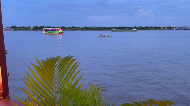 Evening view of the river in Phnom Penh, Cambodia, showing a brightly decorated tourist boat with colorful neon lights alongside a smaller canoe carrying locals