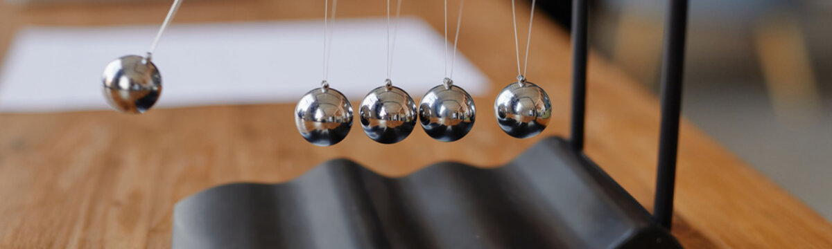 Header of Newton cradle balls swinging on metal frame, close up showing motion and physics concept, reflective metal spheres suspended by strings, wooden table surface visible in background
