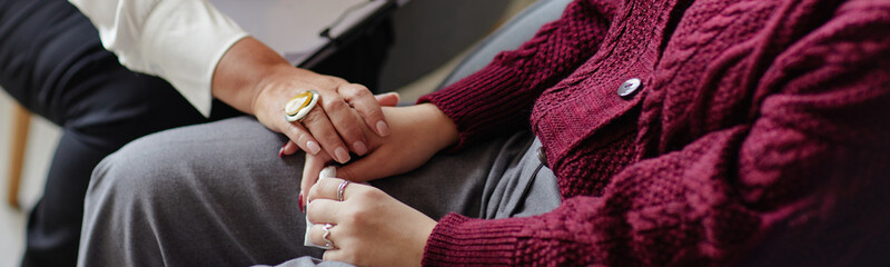 Header of woman comforting young adult woman by holding hands, both seated closely together,...