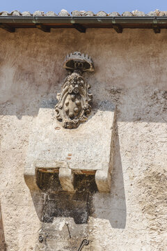 View of a textured stone wall adorned with an intricate coat of arms and corbel casting shadows against the sunlit facade, Santo Stefano di Sessanio, Abruzzo, Italy.
