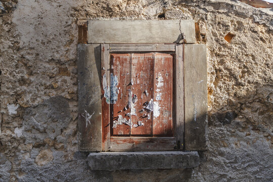 View of a weathered, muted red, boarded-up window, marked by peeling posters and framed by crumbling stone and concrete textures, Calascio, Abruzzo, Italy.