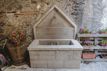 View of an aged stone fountain, adorned with potted plants, stands against a textured wall, a rustic scene evoking timeless charm, Castelvecchio Calvisio, Abruzzo, Italy.