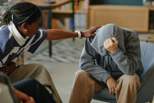 Black man comforting distressed teenager sitting with head covered by hood, hand on forehead, supportive gesture in indoor setting, emotional support and empathy visible