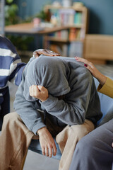 Teenager boy with hood covering head sitting with face in hands, receiving comfort from multiethnic...