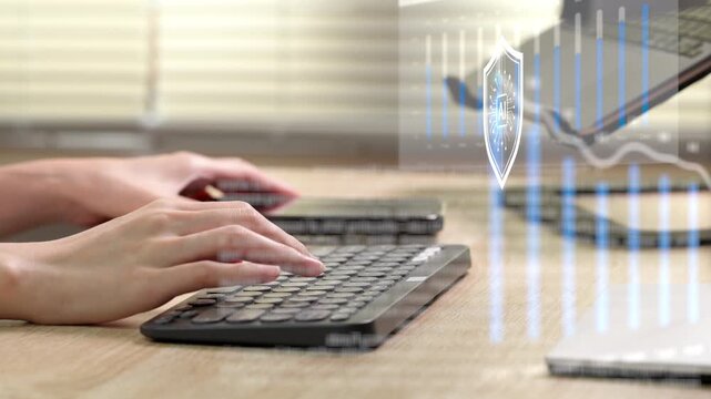 A person in a white shirt works on a laptop placed on a wooden desk beside window blinds in a home office.