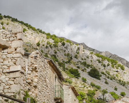 View of the ancient stone buildings nestled against a dramatic, rocky hillside, under a cloudy sky in Frattura Vecchia, Abruzzo, Italy.