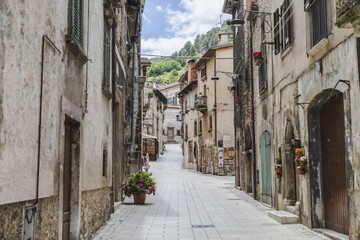 View of aged stone buildings line a narrow street with flower pots, under a bright sky, leading to distant green hills, Scanno, Abruzzo, Italy.