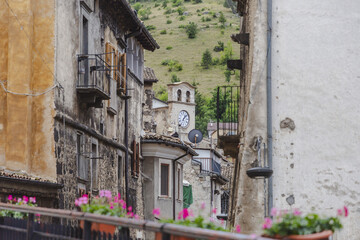 View of a weathered clock tower rising above aged buildings with pink flowers in the foreground, set against a green hillside, Scanno, Abruzzo, Italy.