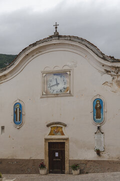 View of an old church facade, adorned with a clock and religious art, stands weathered yet dignified against a muted sky, Castrovalva, Abruzzo, Italy.