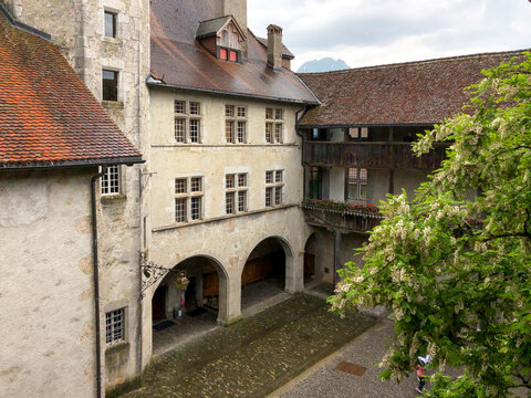 View of weathered stone walls embrace a courtyard, sunlight filtering through arched walkways and blooming trees, creating a serene atmosphere, Gruy&Atilde;&uml;res, Fribourg, Switzerland.