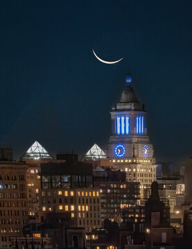 View of the moon hangs like a silver scythe over illuminated buildings, casting a cool glow on the urban scene, New York, New York, United States.