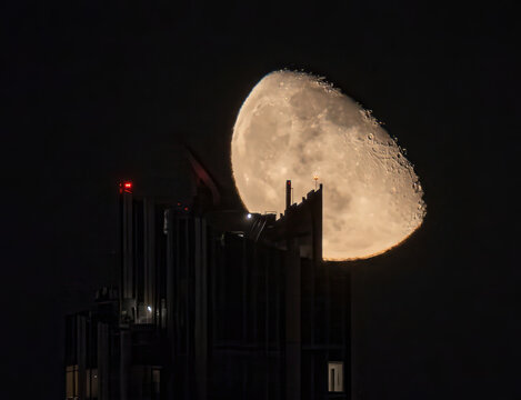 View of the moon seemingly perched atop a skyscraper, casting a warm glow against the dark sky, a surreal urban celestial dance, New York, New York, United States.